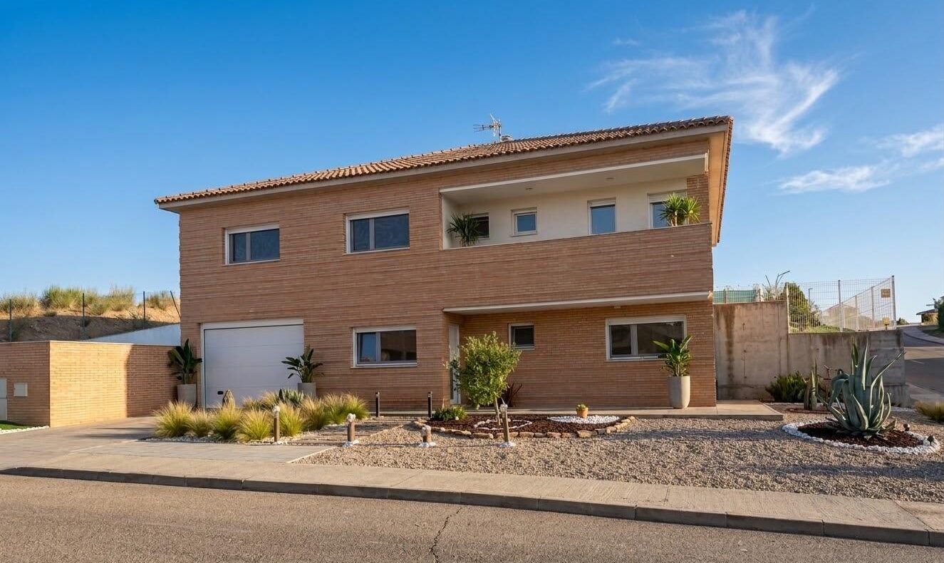 Two-story brick house with a flat front yard, a white garage door, and desert landscaping under a clear blue sky.