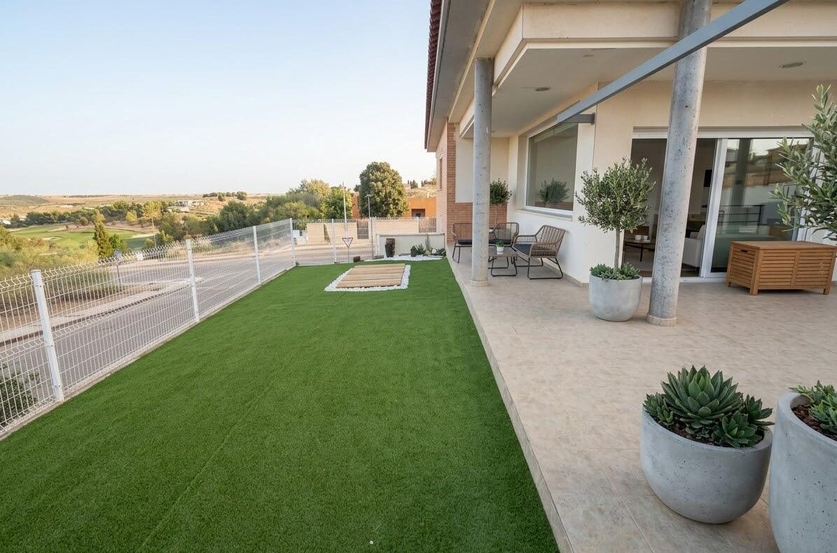 Terrace with beige tiled patio and artificial grass, potted plants, and outdoor seating along a glass door, overlooking a rural landscape.