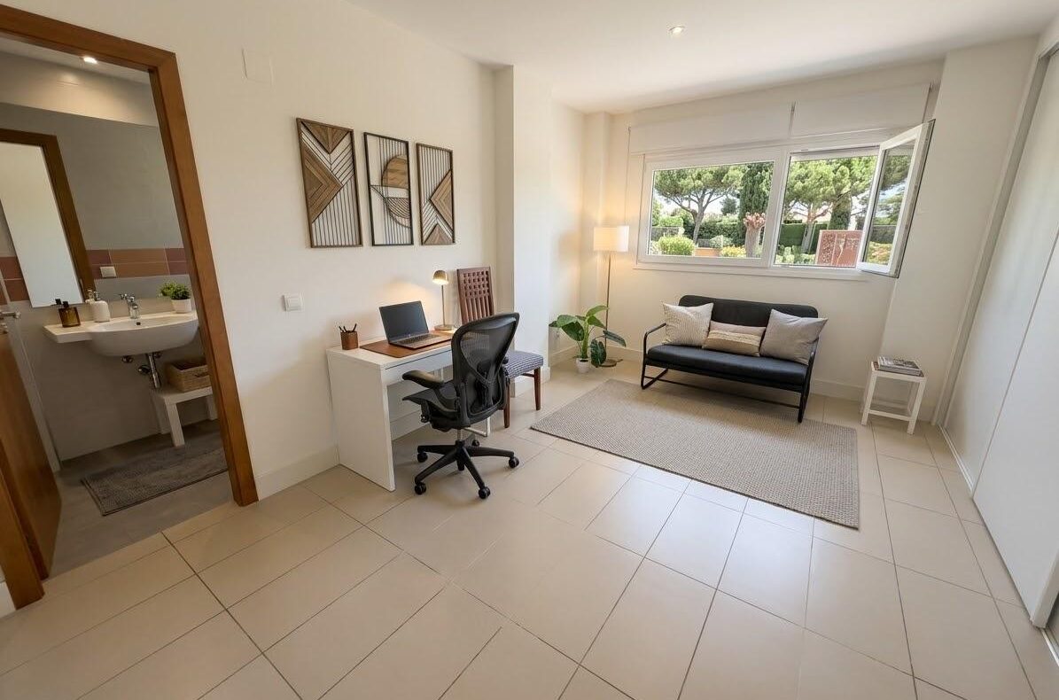 Bright home office and small sitting area: white desk with laptop and ergonomic chair, facing a living area with a black loveseat, rug, and large window showing greenery outside.