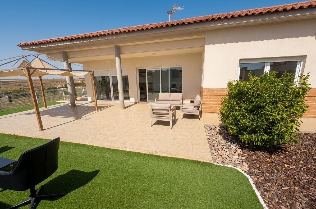 Beige tiled patio of a single-story home with outdoor seating and sliding glass doors, under a clear blue sky.