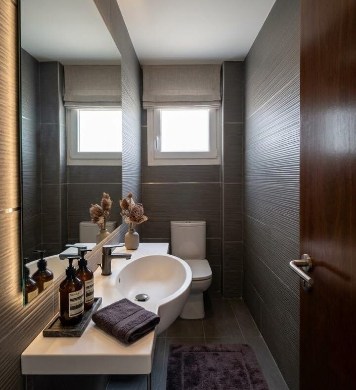 Modern bathroom with a white oval sink, dark tile walls, mirror, and a toilet at the far end; purple towels and a vase of dried flowers on the counter.