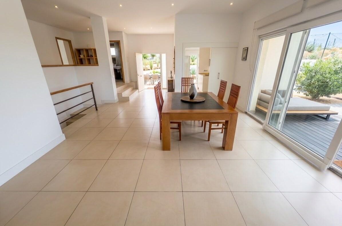 Bright open dining area with a wooden table and six chairs, beige tiled floor, and large sliding glass doors to a patio.