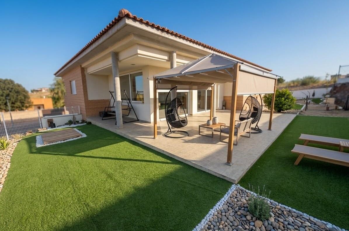 Backyard view of a modern house with a covered patio, hanging chairs, and a green artificial lawn area surrounded by pebbles and planters.