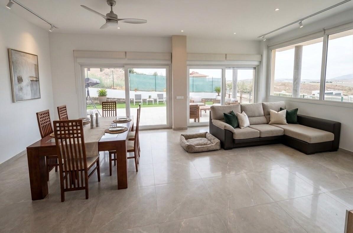 Bright open-plan living and dining area with sliding glass doors to a patio, a beige tiled floor, and a large L-shaped sofa.