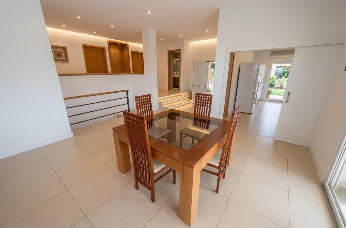 Open-plan dining area with a wooden square table, four lattice-back chairs, and a glass top; bright, tiled floor and garden view beyond.