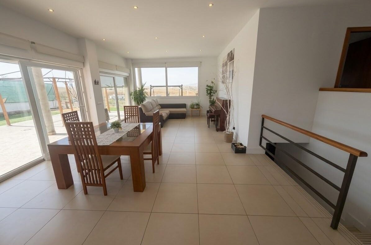 Bright open-plan dining and living area with a wooden table, six chairs, beige sofa, and large glass doors to the outside patio.