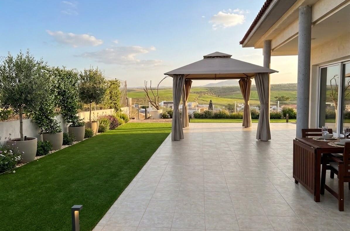 Patio with a beige canopy gazebo on a tiled deck, expansive lawn, and outdoor dining area with a wooden table to the right, open fields in the distance.