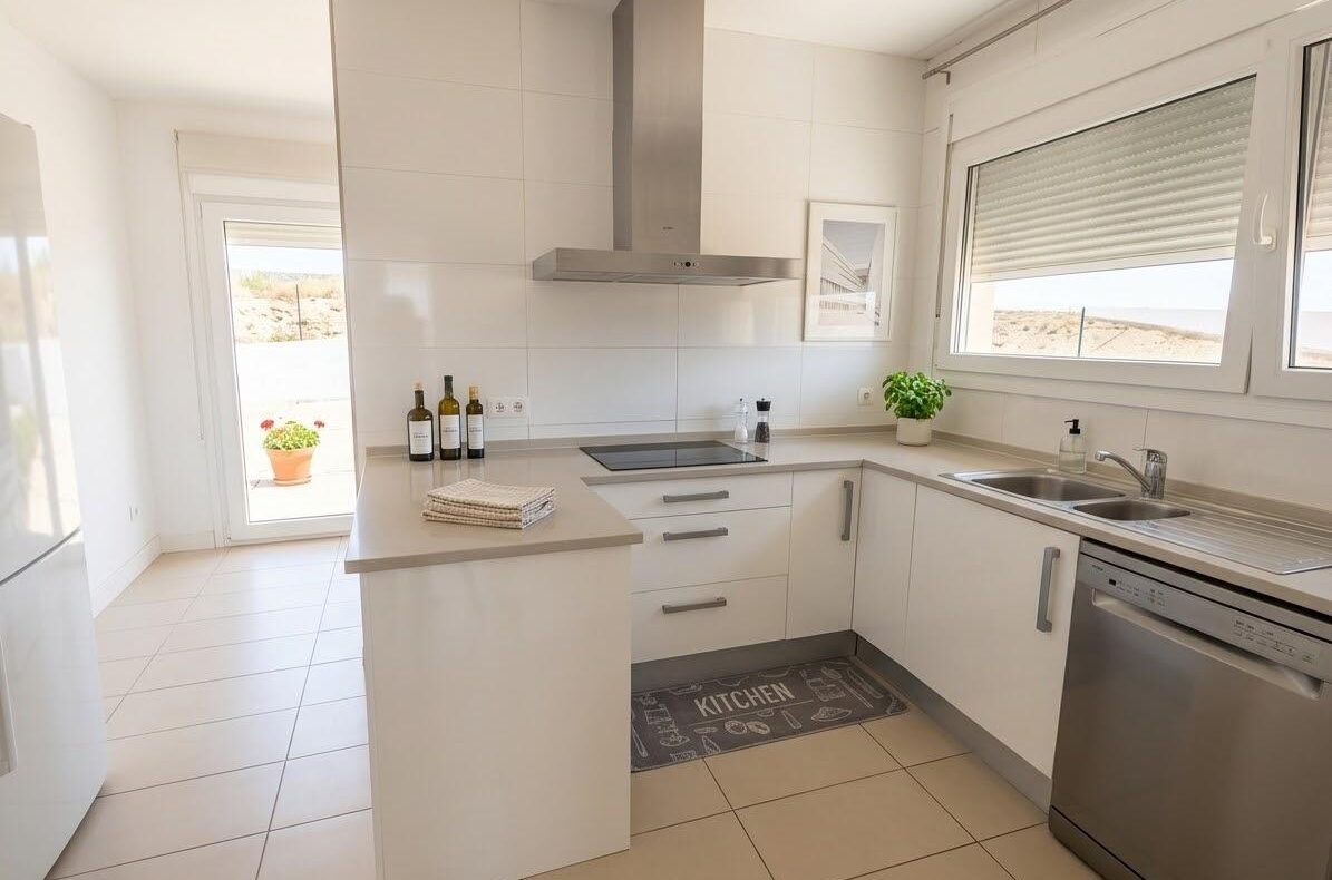 Bright white modern kitchen with an island, stainless steel range hood, and a windowed sink area; bottles on the counter and a potted plant nearby.