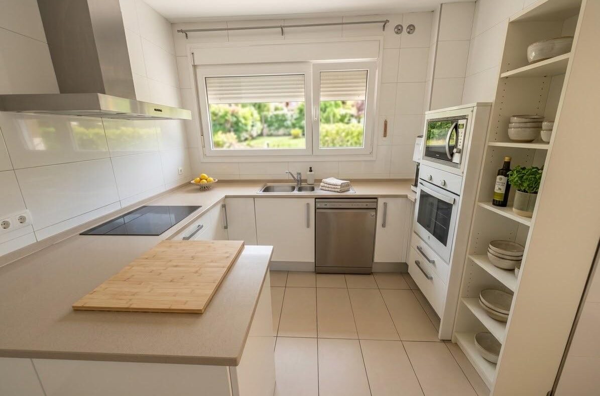 Bright white kitchen with beige countertops, an island and cutting board, window over the sink, and stainless steel appliances.
