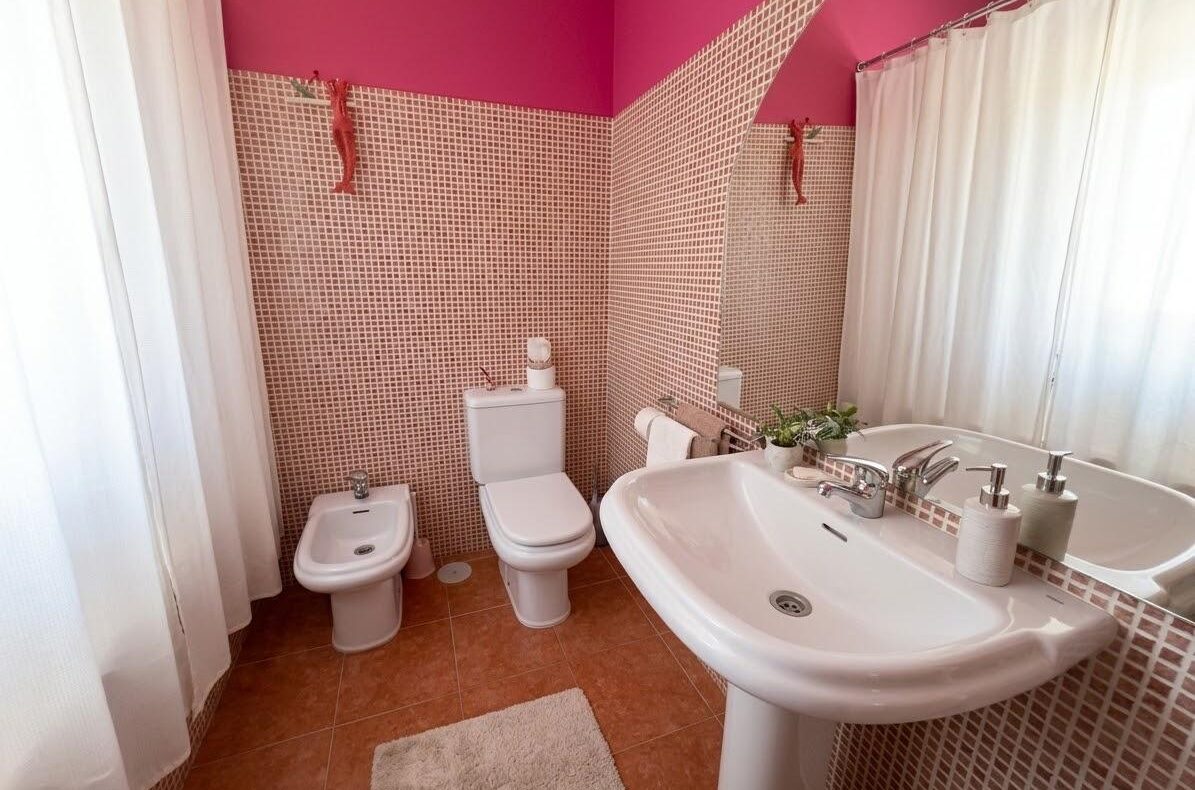 Bright pink-tiled bathroom with a white pedestal sink, toilet, and bidet, and a white shower curtain along the tub wall.