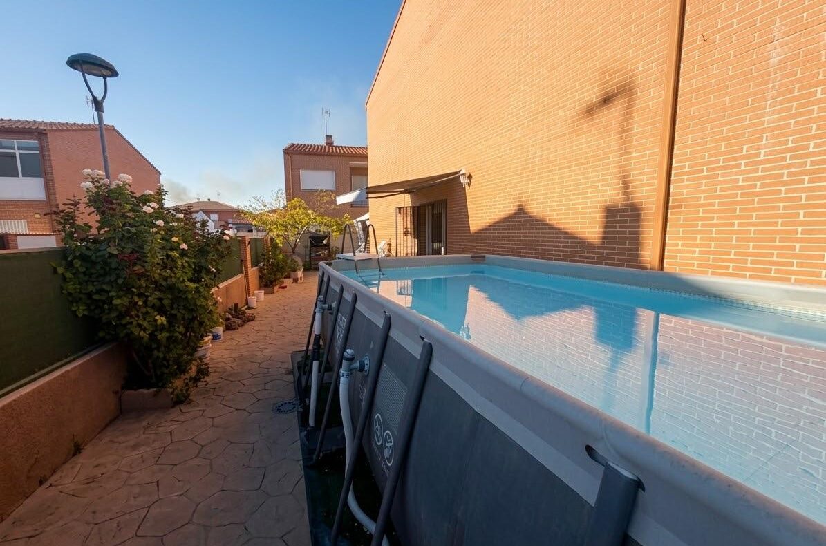 Above-ground swimming pool beside a brick house, with a stone pathway and potted plants lining a green fence on the left under a clear blue sky.