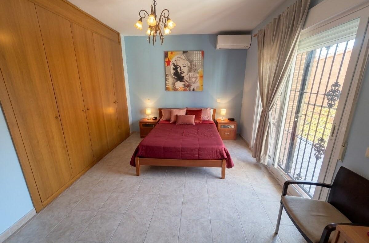 Bedroom with a wooden-framed bed on a blue wall, magenta bedding, two lamps, and a large built-in wooden wardrobe to the left.