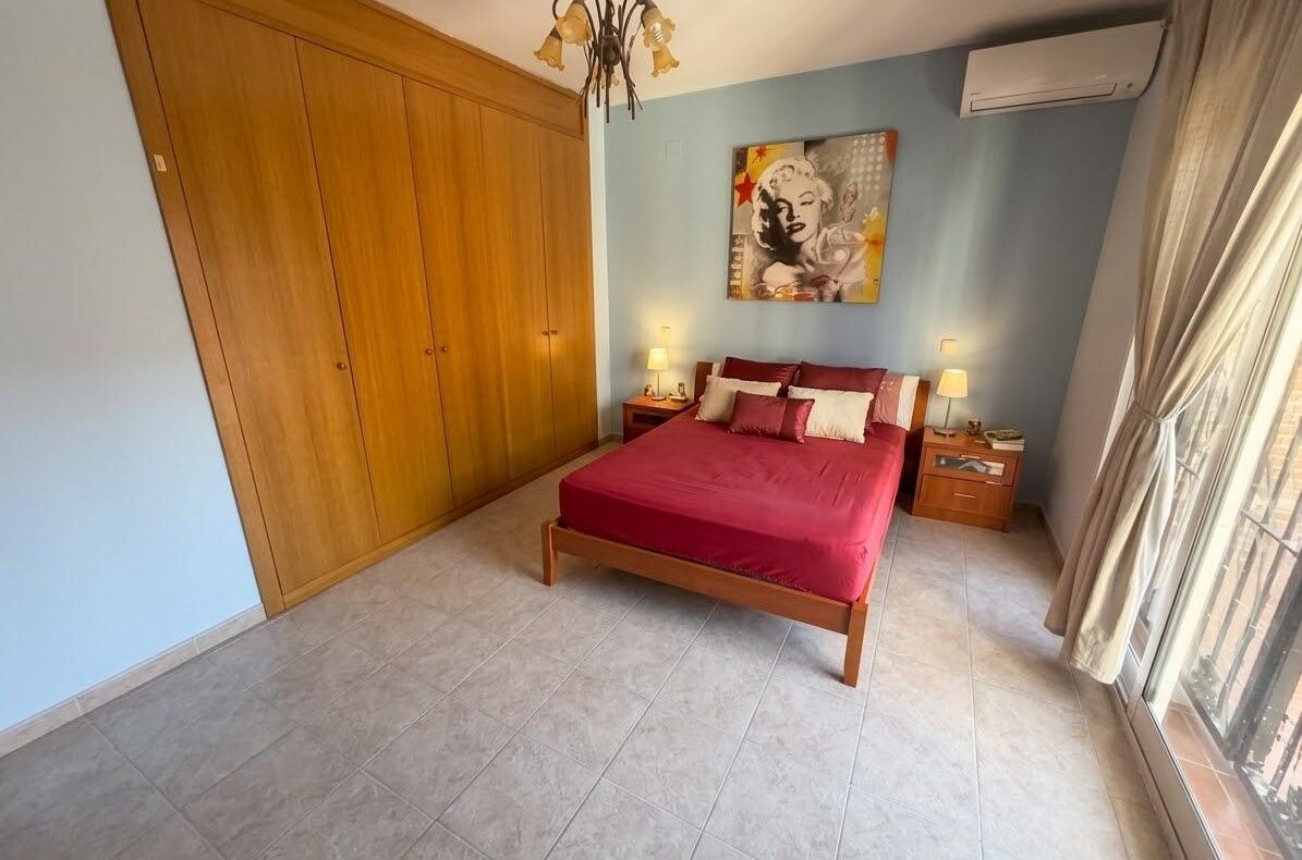 Bedroom with a red bedspread, large wooden wardrobe, two nightstands, and a Marilyn Monroe artwork on a blue wall.