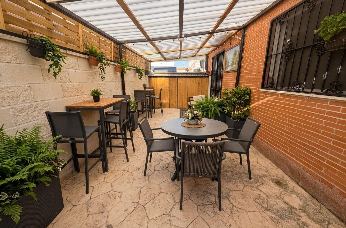 Outdoor patio with hexagonal stone tiles, black metal dining set and tall bar tables, potted plants along a brick wall under a translucent roof.