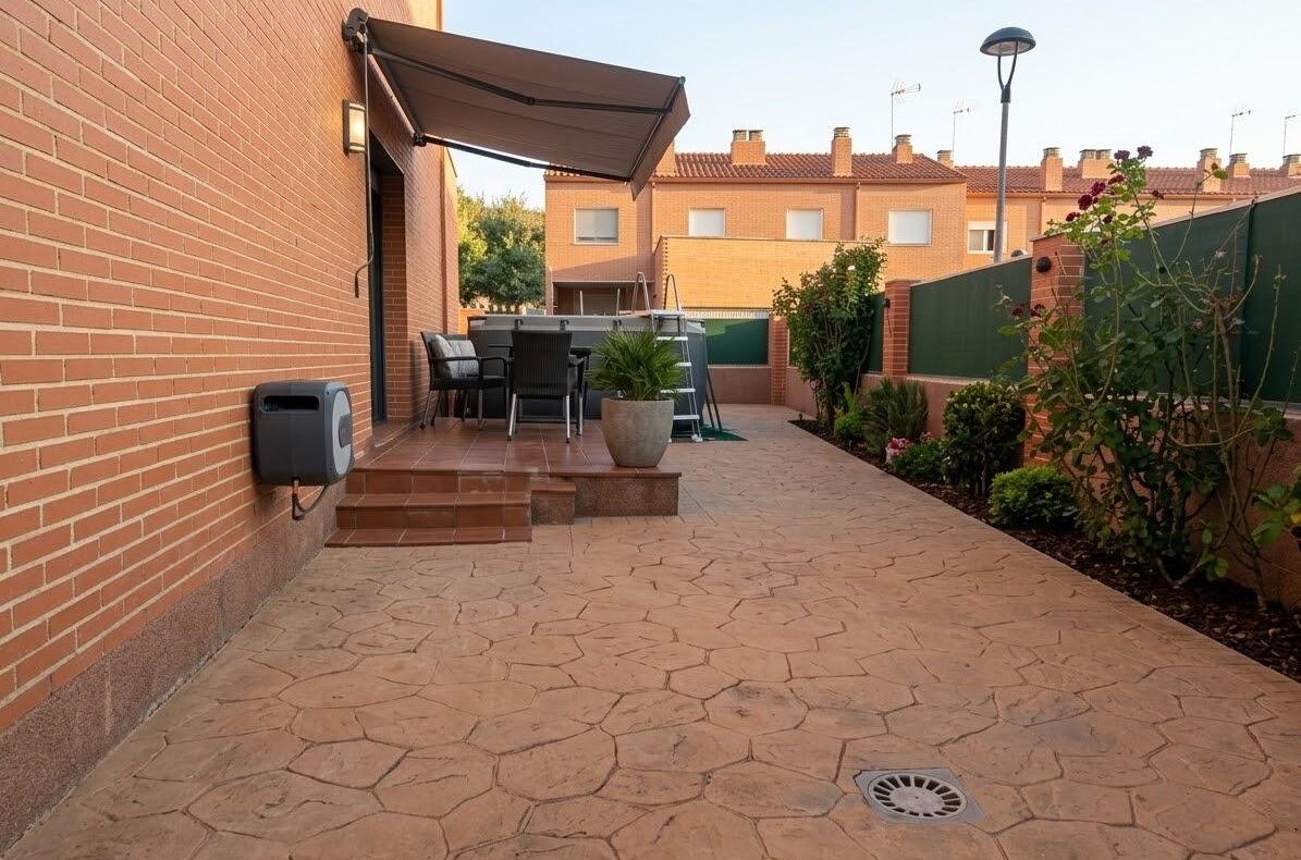 Patio area beside a brick house with a retractable awning, outdoor dining table and chairs, potted plant, and stone-patterned paving on a sunny day