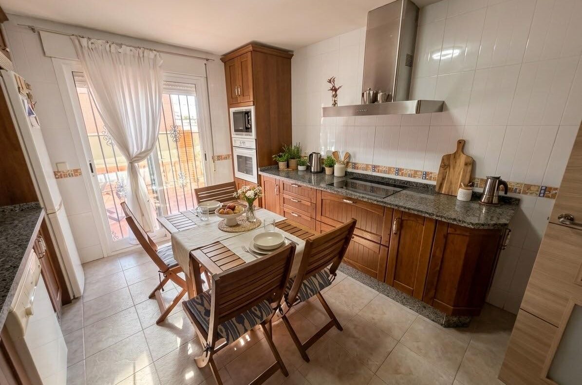 Bright kitchen with wooden cabinets, granite countertop, and a four-seat dining table by a glass door with sheer white curtains.