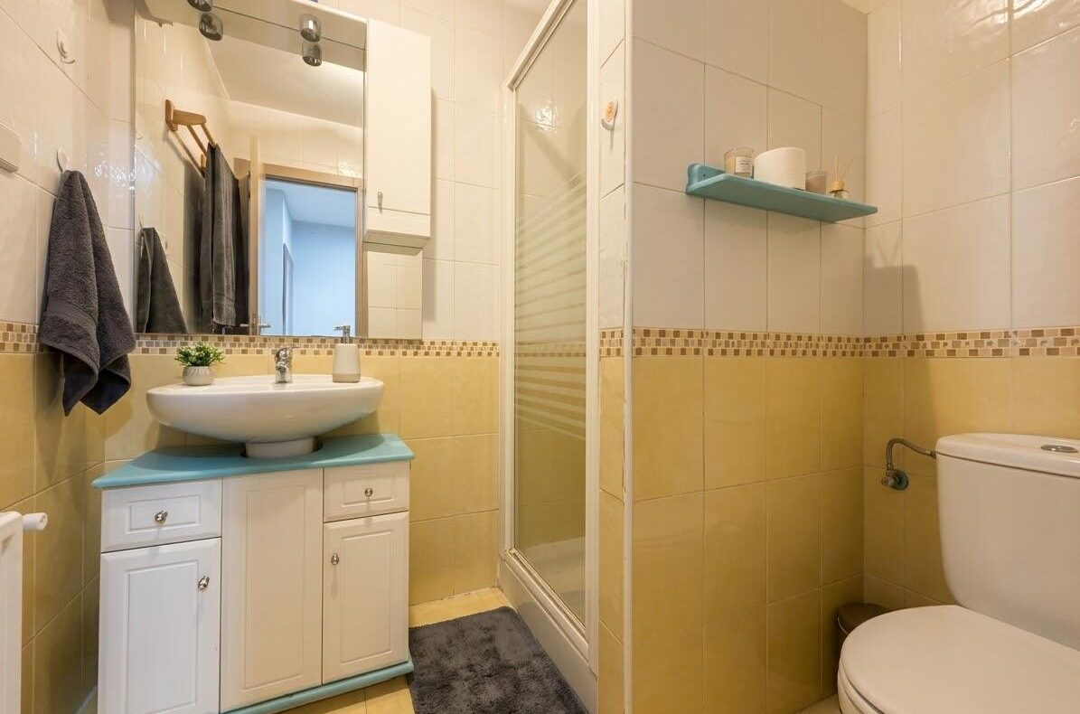 Compact bathroom showing a white vanity with a blue countertop, mirror, and gray towels on a rack beside it; shower to the right and a toilet to the far right; yellow-tiled walls with a decorative border.