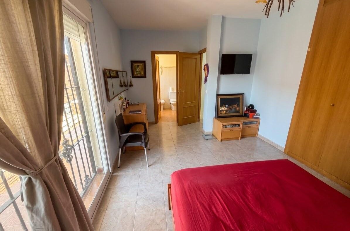 Bedroom with red bedspread, blue walls, wooden wardrobe, and a wall-mounted TV; small desk and chair near a sliding glass door to a balcony, with an open doorway to a bathroom visible.