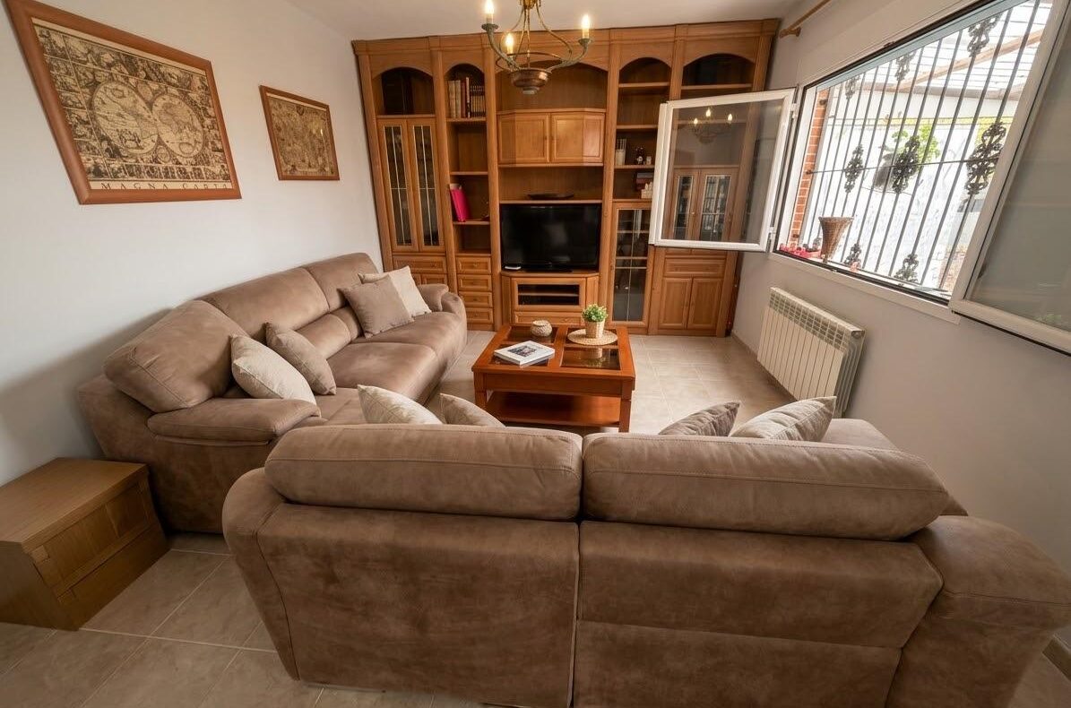Cozy living room with a large L-shaped beige sofa facing a wooden entertainment unit with TV, glass coffee table, and chandelier above.