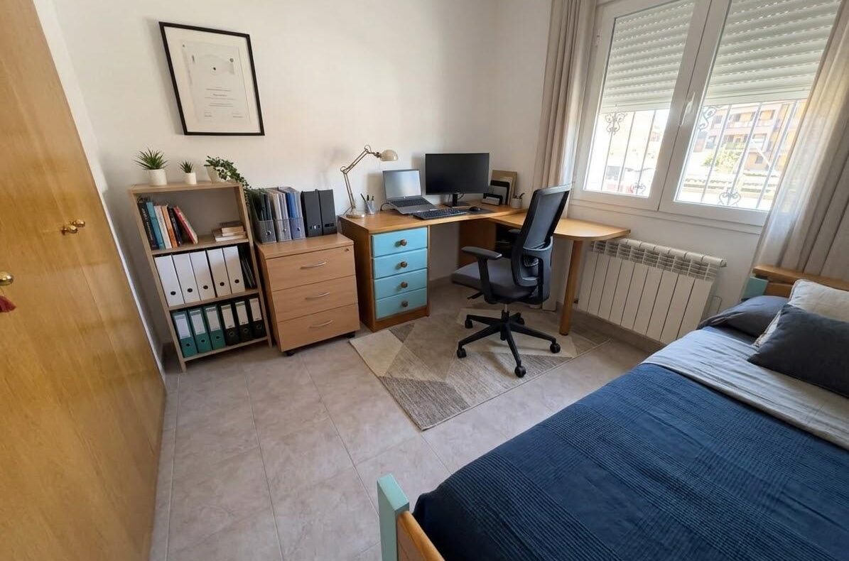 Bedroom workspace: desk with computer monitor and laptop, office chair, and a filing cabinet next to a bookshelf with binders.